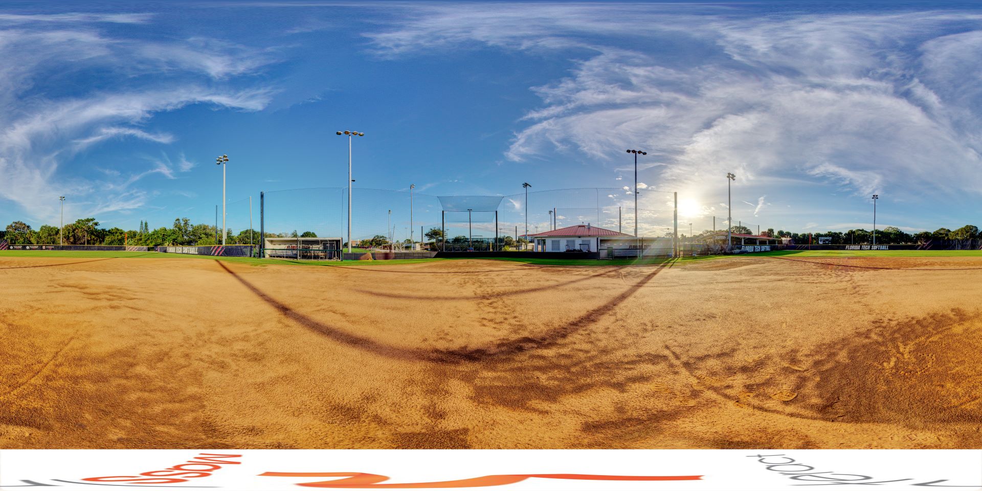 A panoramic view of Nancy Bottge Field at Florida Tech, showing the softball field with its brown dirt infield, green outfield, and various buildings and equipment under a clear blue sky with some clouds